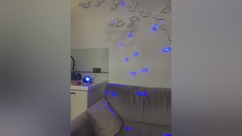 Media: Video of a modern living room with a beige couch, minimalist white wall decor featuring leaf shapes with blue lighting, and a compact kitchenette with white cabinets.