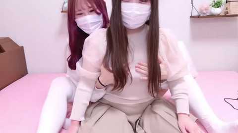 Media: Video of two young Asian women in surgical masks, white scrubs, and beige pants, seated closely on a pink bed. The woman in front holds the other's breasts. Background includes white walls, shelves, and a brown cardboard box.