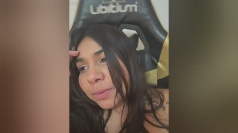 Video of a Latina woman with long, dark hair and a light skin tone, wearing a black tank top, lying on a black gaming chair with a yellow and white logo.