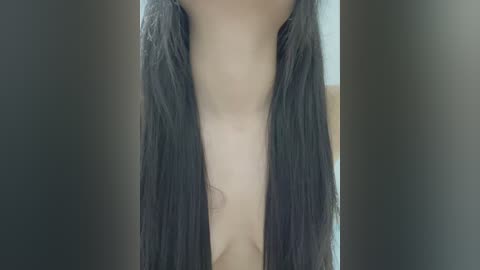 Media: Video of a person with long, straight black hair parted down the center, exposing their bare chest and smooth, light skin. The background is blurred, focusing attention on the subject's torso.