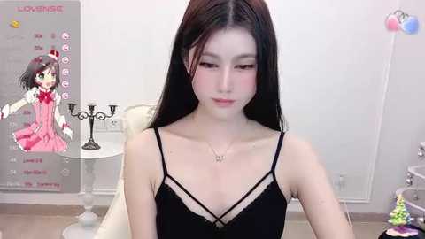 A video of a young Asian woman with long black hair and fair skin, wearing a black lace dress, sitting in a modern room with a poster of a character on the wall.