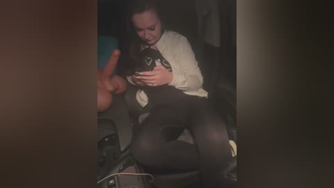 Media: A video captures a woman in a dimly lit room, seated on a black chair, wearing a white blouse and black tights, holding a black handbag.