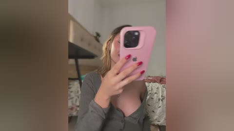 Media: A video of a young woman with fair skin and blonde hair, taking a selfie in a bedroom. She wears a gray cardigan and has red-painted nails. Background includes a bed with floral bedding and a beige dresser.