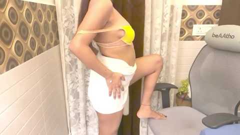 Media: Video of a slender, light-skinned woman in a yellow bikini, adjusting her white skirt, standing next to a gray office chair in a tiled room with patterned walls.