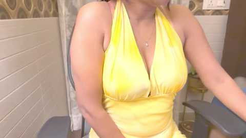 Media: Video of a curvy, light-skinned woman in a yellow halter dress, sitting on a blue chair in a tiled room with a floral patterned wall.