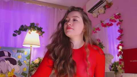 Media: Video of a young Caucasian woman with long, wavy brown hair, wearing a sheer red top, standing in a vibrant, floral-decorated room with a white lamp, framed art, and air conditioner.