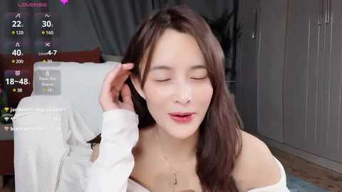 Media: Video of a young Asian woman with long brown hair, wearing a white off-shoulder top, smiling, adjusting her earpiece, in a modern living room with grey cabinets.