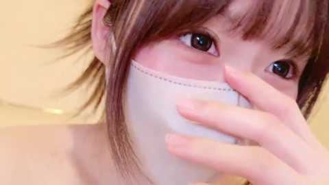 Media: A close-up video of a young Asian woman with brown hair and dark eyes, wearing a white surgical mask, with a blurred, pale yellow background.