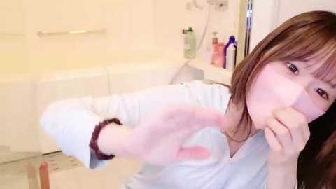 Media: A video of a young Asian woman with brown hair, wearing a white shirt, making a \"stop\" gesture with her hand in a bathroom with beige walls, a bathtub, and toiletries on the ledge.