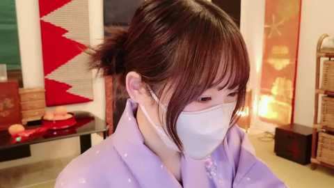 Media: Video of an Asian woman with long brown hair, wearing a white mask and light purple robe, in a brightly lit room with traditional Japanese art and decor.