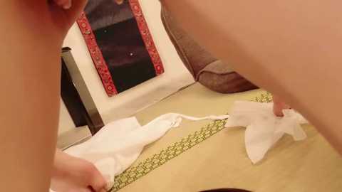 Media: Video of a person, likely a woman, with fair skin, wearing white panties, removing a white garment from a yellow and green-patterned mat. Background includes a red-framed mirror and a red-patterned wall.