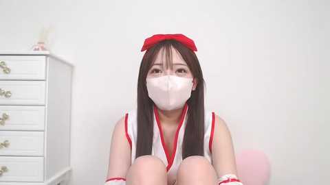 Media: Video of an East Asian woman with long black hair and fair skin, wearing a white face mask, red headband, and revealing red and white cosplay outfit, sitting on a white chair in a minimalistic white room.