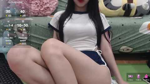 Media: A video of a young Asian woman with long black hair, wearing a white sports shirt and dark blue shorts, sitting on a green couch in a cozy room with a gaming monitor showing a virtual reality game.