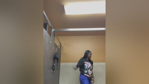 Media: Video of a young Black woman with long braids in a public bathroom stall, wearing a black graphic T-shirt and blue pants. The stall has beige walls and a fluorescent light overhead.