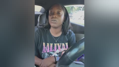 Media: A video of a young Black woman with shoulder-length black hair, wearing a black \"Malikai\" t-shirt, sitting in a car. She has a neutral expression, and the car's interior is visible with a steering wheel.