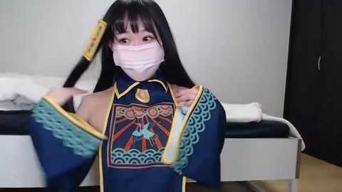 Media: Video of an Asian woman in a traditional Chinese costume, with a mask, sitting on a bed. She has long black hair and is wearing a blue robe with intricate designs. The background is a simple bedroom with white walls and a white bed.