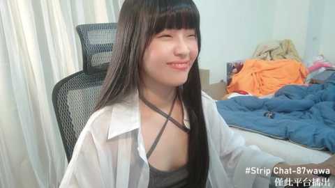 Media: Video of an Asian woman with long black hair and fair skin, wearing a white shirt and grey bralette, smiling. Background shows a messy bed with orange and blue sheets. Text: \"#StripChat #Tawa_Yukihara #Nude-Yukihara\