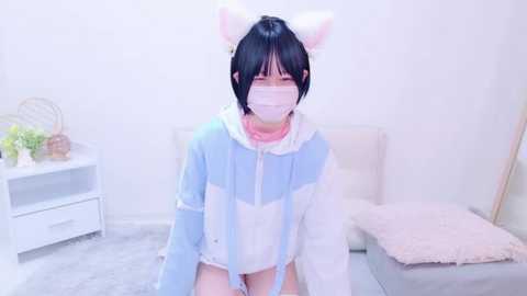 Media: Video of an Asian woman with black hair, wearing a white and blue cat hoodie and cat ears, sitting on a white couch, wearing a mask, in a minimalist white room with a green plant and a pink cushion.