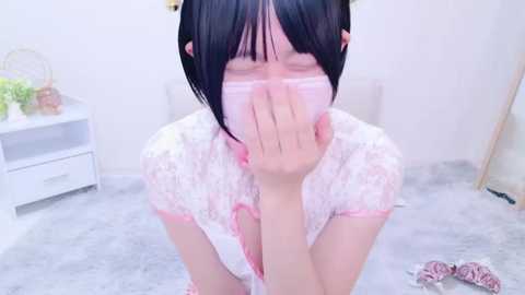 Media: Video of an Asian woman with short black hair, wearing a white lace dress, covering her mouth with her hand, standing on a grey carpeted floor in a simple, white room with a white nightstand and a mirror.