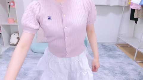 Media: Video of a fair-skinned woman wearing a pink, button-up cardigan over a white, floral-patterned skirt, standing in a pastel-themed room with light-colored furniture and soft lighting.