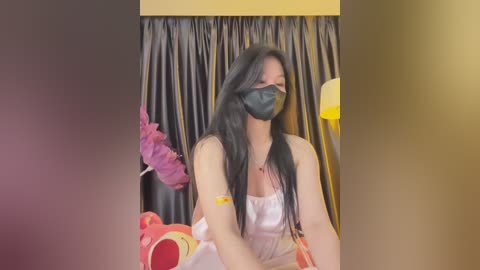 Media: Video of a young woman with long black hair, wearing a black face mask and white dress, seated against a dark curtain backdrop, holding a pink feather duster and a plush toy.