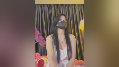 Media: Video of an Asian woman with long black hair, wearing a black face mask, white tank top, and yellow bracelet, seated in front of a pink teddy bear and a dark curtain background.