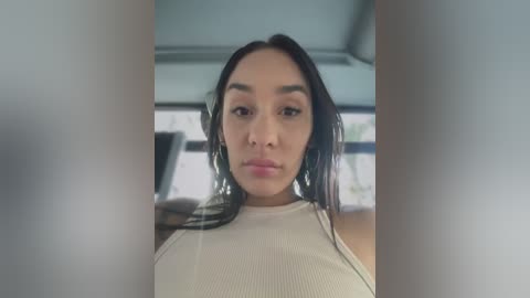 Media: Video of a young woman with medium skin tone, dark hair, and full lips, wearing a light-colored ribbed tank top, taken from inside a car.