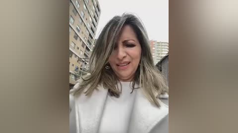 Media: Video of a middle-aged woman with shoulder-length, light brown hair, wearing a white coat, standing in an urban setting with beige and yellow buildings in the background.