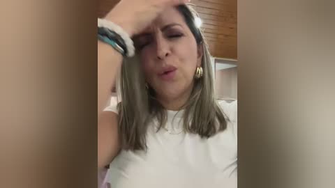 Media: A video of a Caucasian woman with shoulder-length brown hair, wearing a white shirt, rubbing her forehead in a bathroom with wooden ceiling and beige walls.