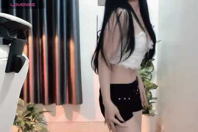 Media: Video of an East Asian woman with long black hair, wearing a white lace crop top and black shorts, standing in a modern, indoor setting with a white wall, a potted plant, and a black curtain in the background.