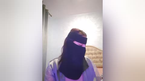 Media: Video of a woman in a light-colored robe, wearing a black mask with pink stripes, standing in a bedroom with a beige headboard and white walls.