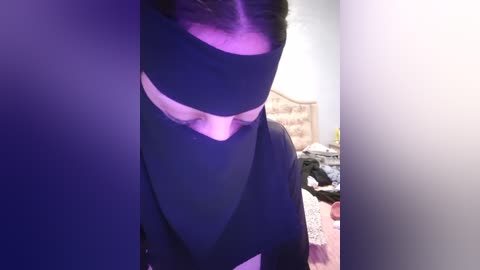 Media: Video of a woman wearing a black burqa in a dimly lit bedroom, her face partially visible, with purple light casting shadows on her headscarf.