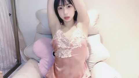 Media: Video of an East Asian woman with fair skin, straight black hair, and red lipstick, wearing a pink satin nightgown with lace trim, lying on a bed with white pillows, arms raised, in a softly lit room.