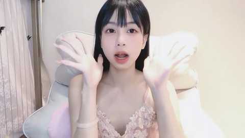 Media: A young East Asian woman with long black hair and fair skin, wearing a pink lace bra, sits on a white chair, with hands raised, in a soft-lit room.