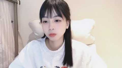 Media: Video of an Asian woman with straight black hair, wearing a white t-shirt, sitting in a chair, with a beige wall and a window with vertical blinds in the background.