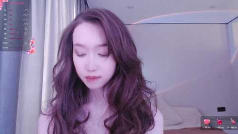 Media: A video of a young Asian woman with long, wavy brown hair, pale skin, and closed eyes, wearing a white top. The background features a bed and a light-colored curtain.