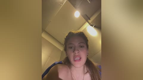 Media: A candid video captures a young woman with light skin, brown hair, and a neutral expression. She's wearing a blue dress. The setting is a dimly lit, beige room with ceiling lights.