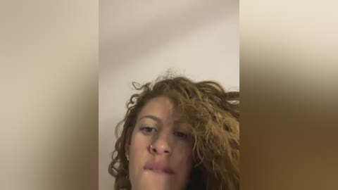 Media: Video of a woman with curly brown hair, light skin, and full lips. The background is a blurred, beige wall. Her expression is neutral, eyes slightly closed, with a hint of a smile.