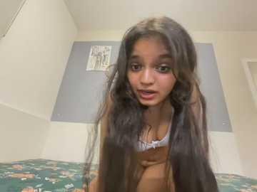 Media: Video of a young South Asian woman with long, dark hair, wearing a white lace bra, standing in a simple, beige-walled room with a colorful floral bedspread.