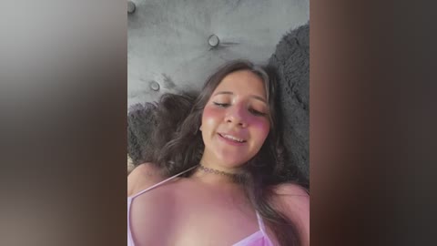 Media: A video of a smiling Latina woman with long dark hair, lying on a tufted gray headboard, wearing a pink lingerie bra, her eyes closed, surrounded by a blurred, dark background.