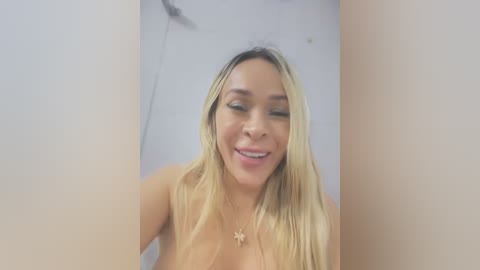 Media: Video of a topless woman with long, blonde hair, light skin, and a small, star-shaped pendant necklace, smiling and winking. She stands in front of a plain, white background.