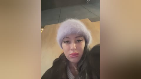 Media: Video of a young woman with pale skin and pink lips, wearing a fluffy lavender beanie and a dark coat, indoors with a wooden floor and a gray ceiling.