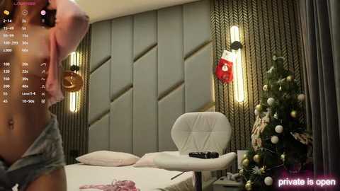 Video of a modern, dimly lit bedroom with a Christmas tree, a white chair, and a bed with a grey headboard. A person is partially visible in the foreground, wearing a pink shirt.