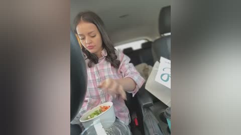 Media: Video of a young girl with shoulder-length dark hair, wearing a pink and white plaid shirt, eating from a white bowl in the backseat of a car.