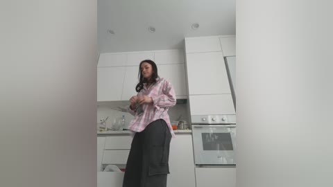 Media: A video of a woman with long black hair wearing a pink plaid shirt and black pants in a minimalist white kitchen with modern cabinets, an oven, and a sink.