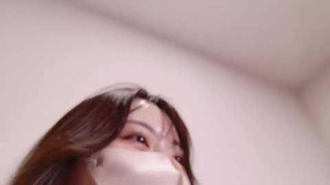 Media: Video of a close-up of a young woman with long, dark brown hair, wearing white makeup, her face partially obscured by a white cloth, in a dimly lit room with off-white walls.