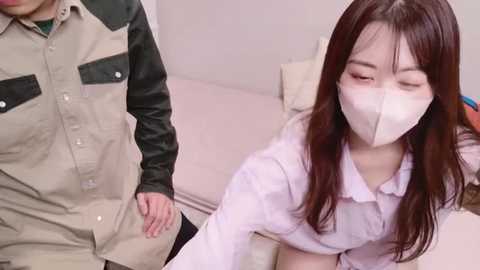 Media: Video of an East Asian woman with long brown hair, wearing a white surgical mask, sitting on a bed, looking down. A man in a beige and black work uniform stands behind her.