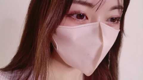 Media: A close-up video of an East Asian woman with long, straight brown hair, wearing a white medical mask that covers her nose and mouth, her eyes gazing slightly downwards.