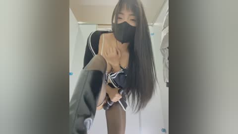 Media: A video of an Asian woman in a black schoolgirl outfit with a black face mask, black knee-high boots, and a black bra, standing in a narrow hallway.