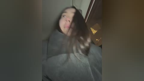 Media: Video of a young woman with long, messy hair, wearing a grey sweater, captured in a dimly lit room with blurry, soft focus.